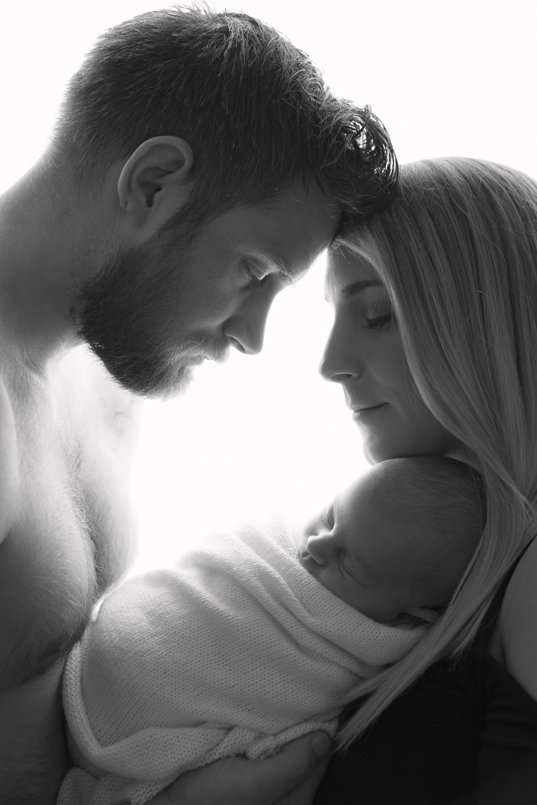 Newborn baby photography by hayley morris photography - newborn photographer - image shows a backlit black and white photo of two parents facing in towards each other and holding their wrapped baby lay between them